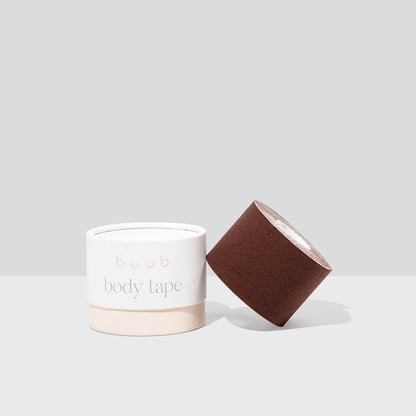 Studio product shot of Buub Body Tape in a deep brown shade. The standard width roll of skin-friendly breast lift tape rests against its white and beige container on a light grey background.