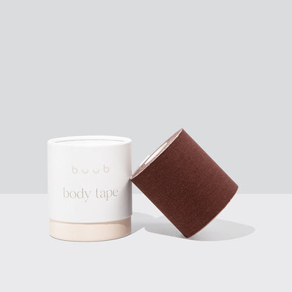 Buub Maxi Body Tape in a deep brown shade for darker skin tones. The wide adhesive boob tape roll is propped against its white and beige container.