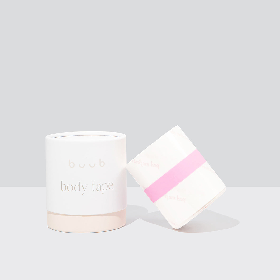 A roll of Buub Ultra Clear Ultra Body Tape with a pink centre strip, shown next to its signature white and beige cylindrical packaging. A discreet breast tape solution for versatile support.
