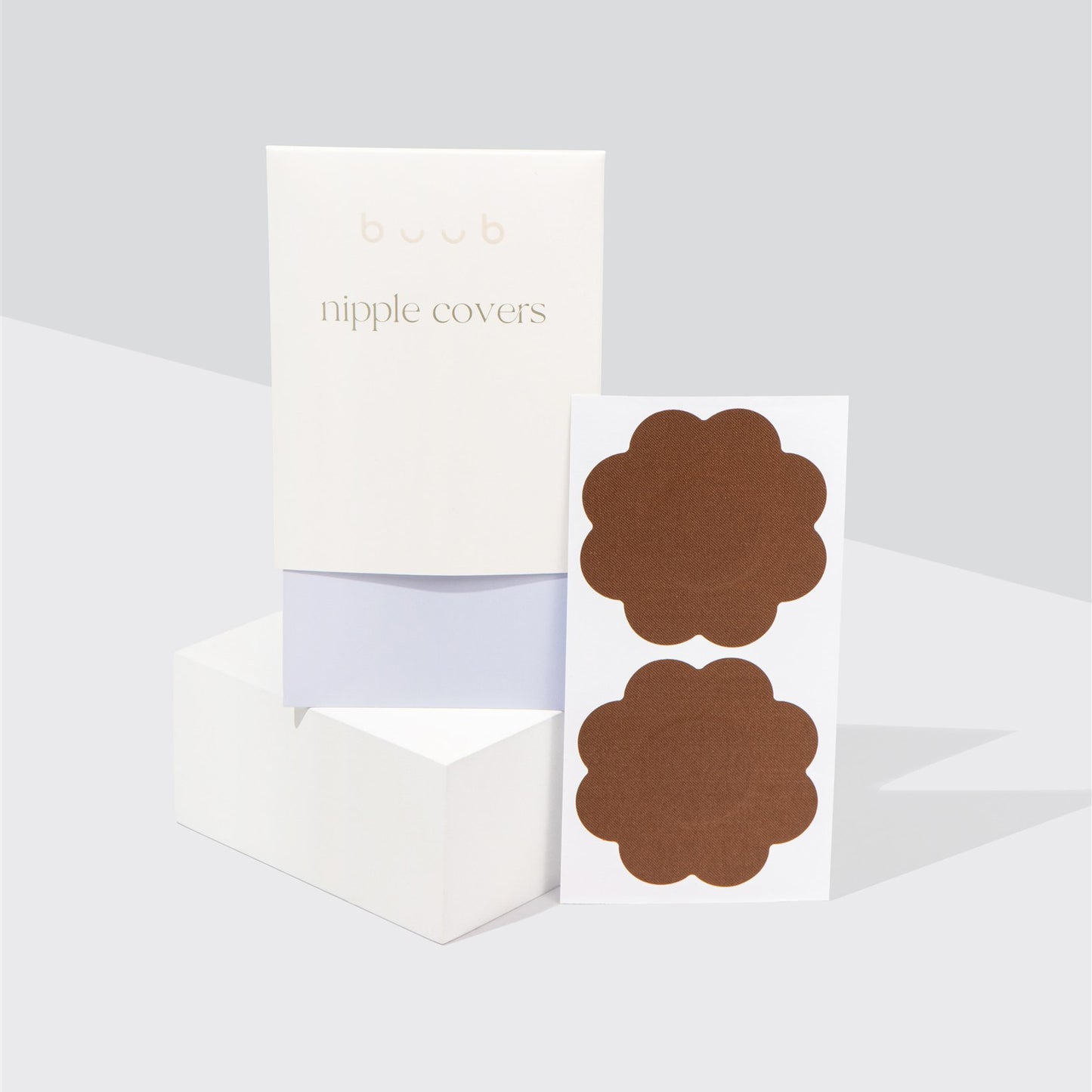 A pair of Buub fabric nipple covers in a deep brown shade, suitable for darker skin tones. The flower-shaped disposable pasties are shown with their chic white and lilac packaging.