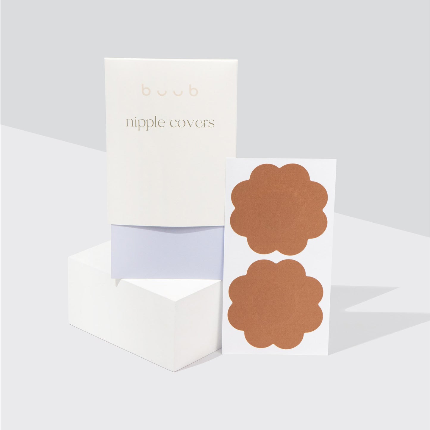 A pair of Buub fabric nipple covers in a versatile tan shade. The disposable flower-shaped pasties are displayed on their backing paper next to their white and lilac branded packaging.