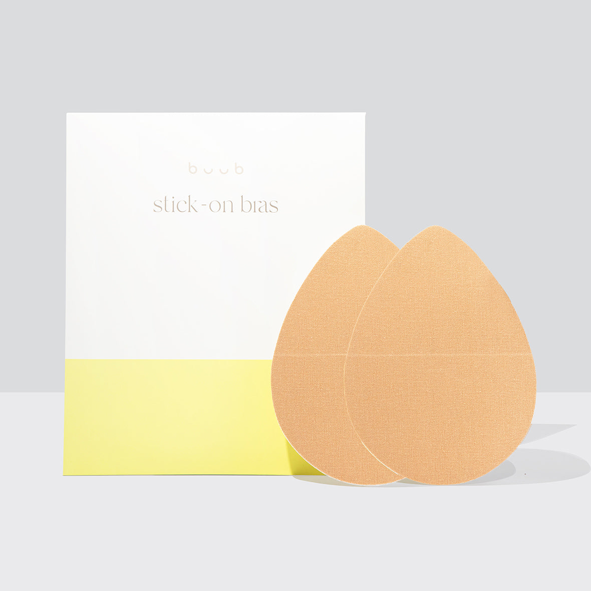 A pair of Buub Stick-On Bras in a nude fabric, providing lift and support. The teardrop-shaped adhesive bra is shown in front of its modern white and yellow packaging.
